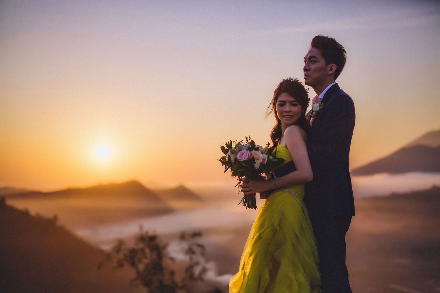A couple in elegant attire embracing during golden hour with mountains in the background.
