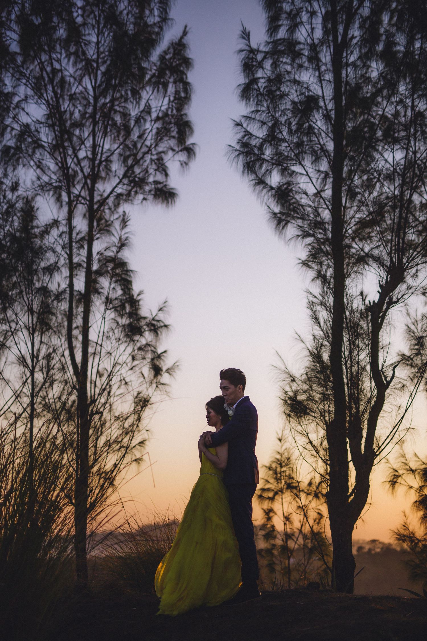A couple in formal wear standing among tall trees at dusk.