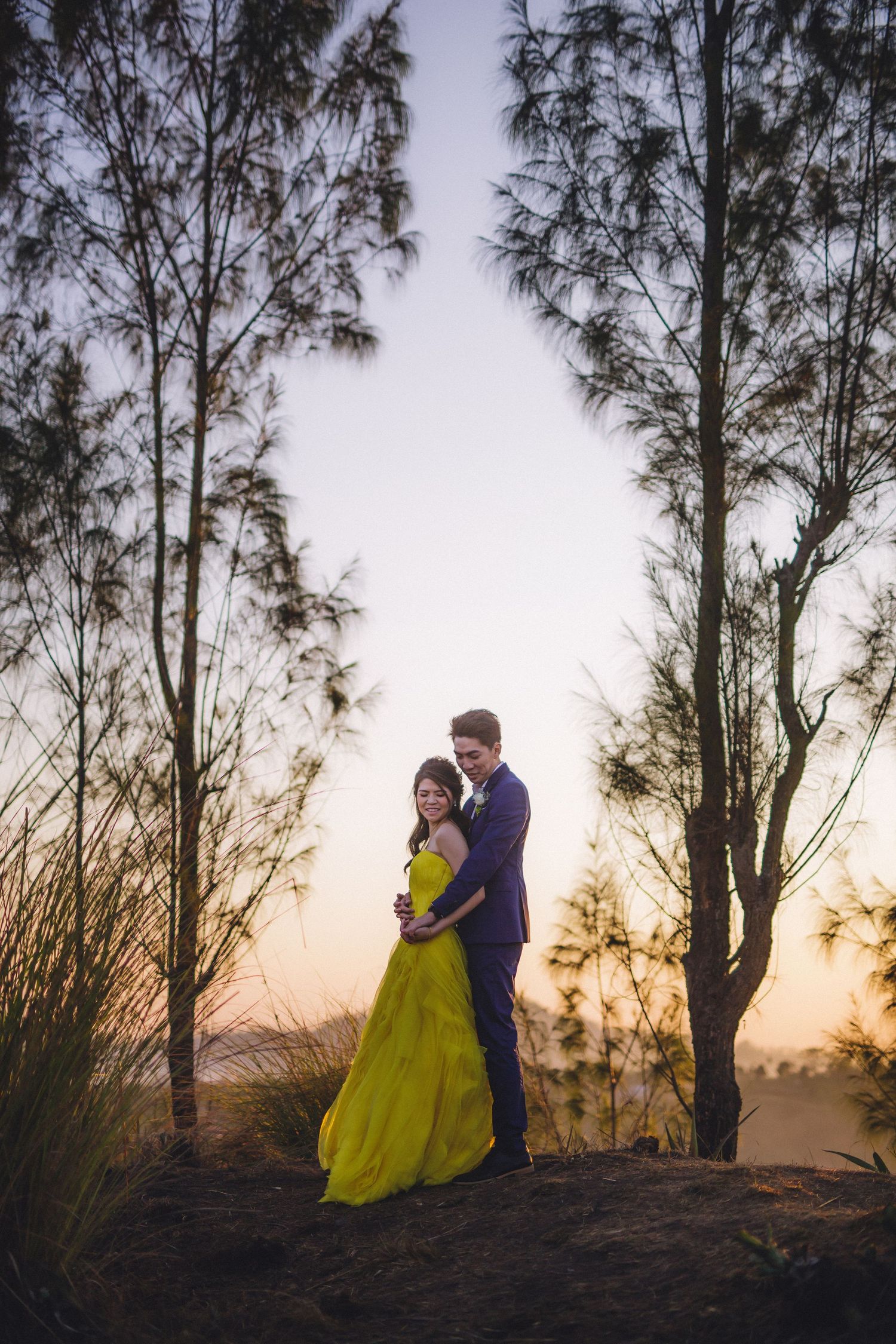 Two people in formal attire posing between tall trees during sunset.