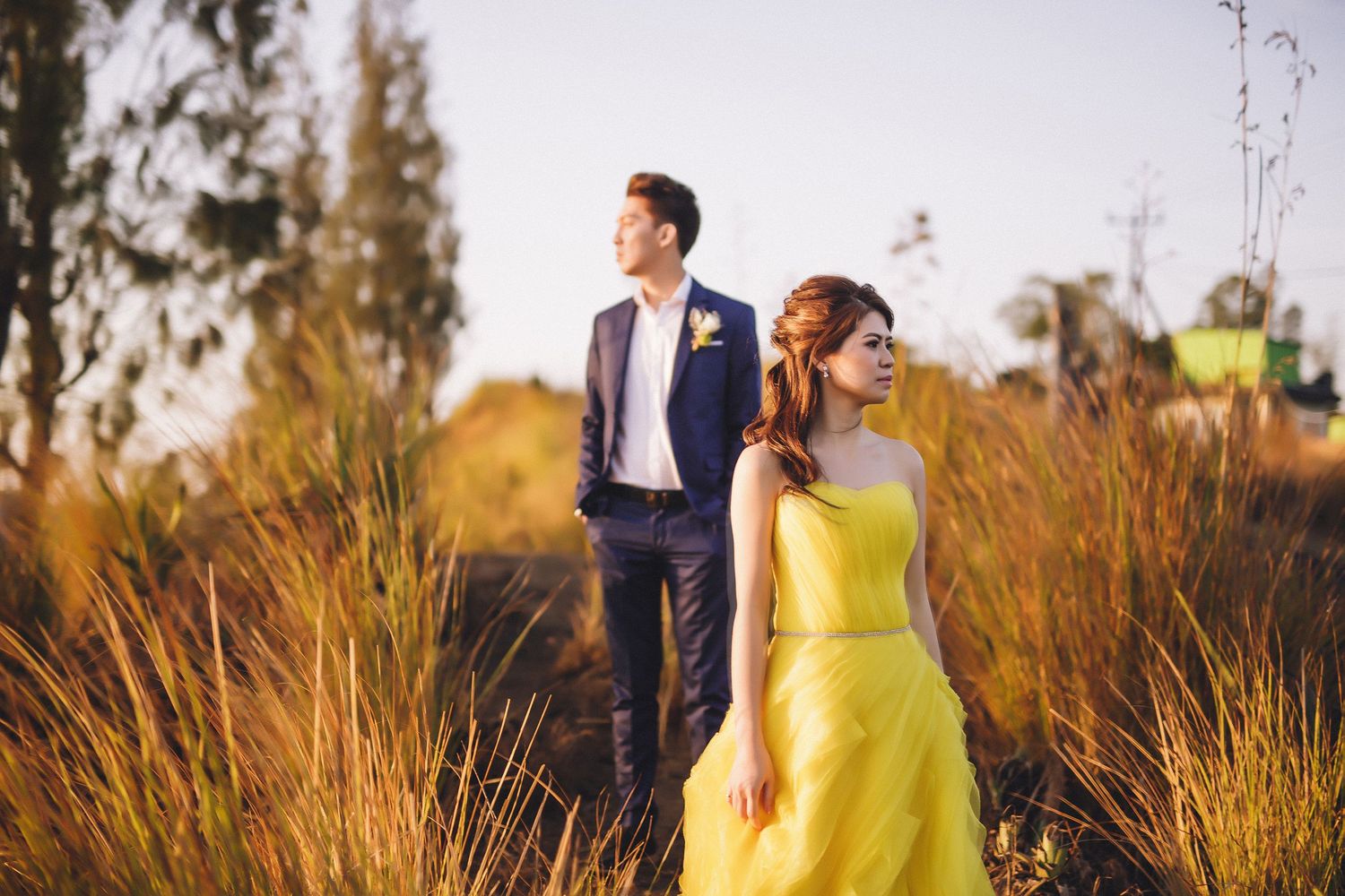 Couple in formal attire walking through golden grass at sunset.