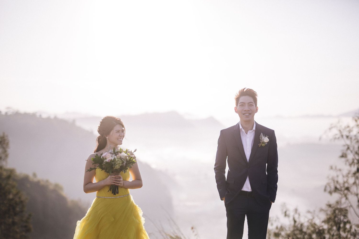 A couple in formal attire stands apart on a misty mountain overlook.