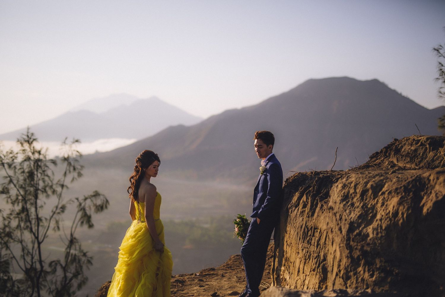 A couple in yellow dress and navy suit stands on a cliff edge with mountain views.