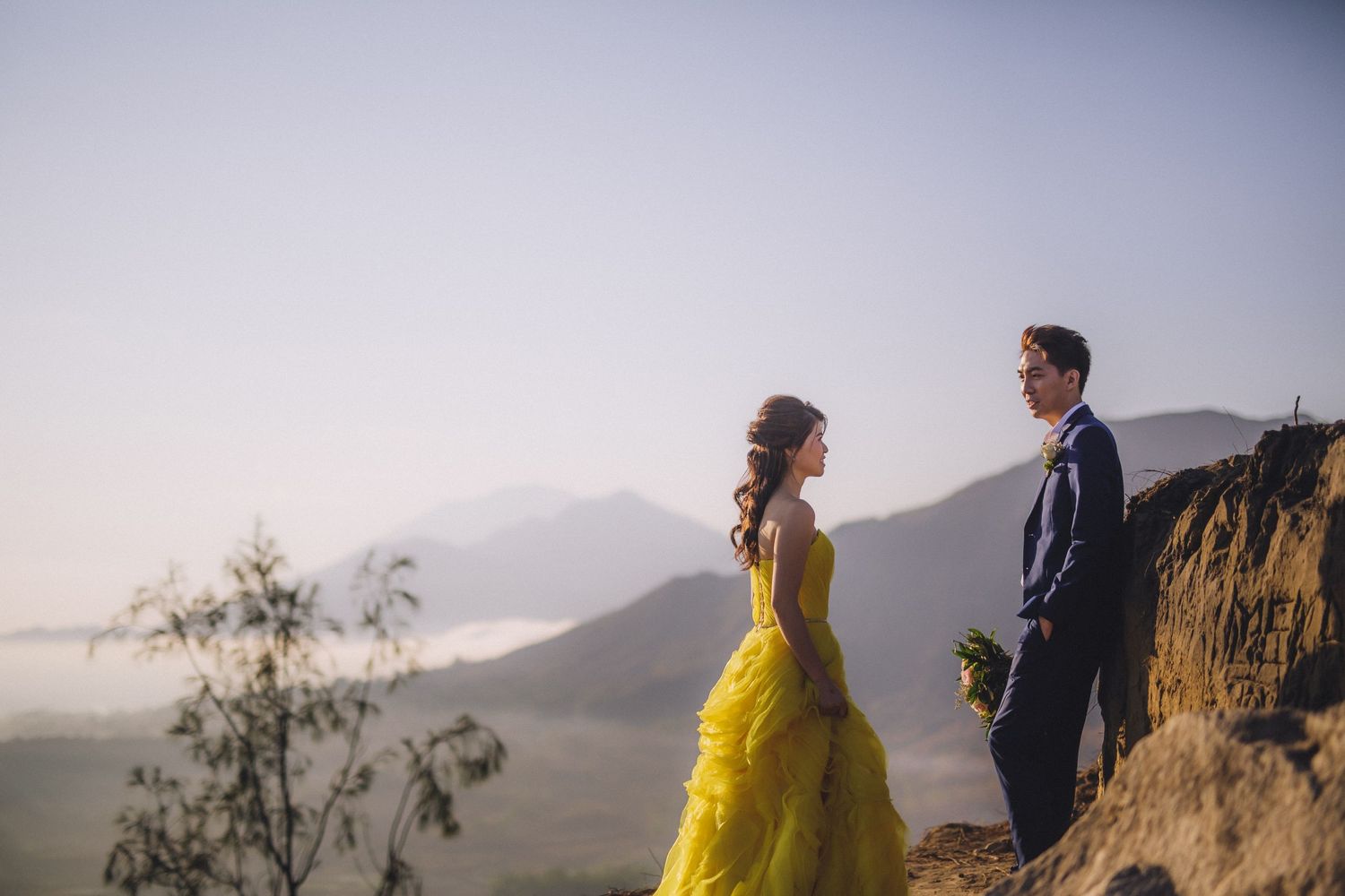A couple in formal wear shares a moment on a mountain cliff during sunset.