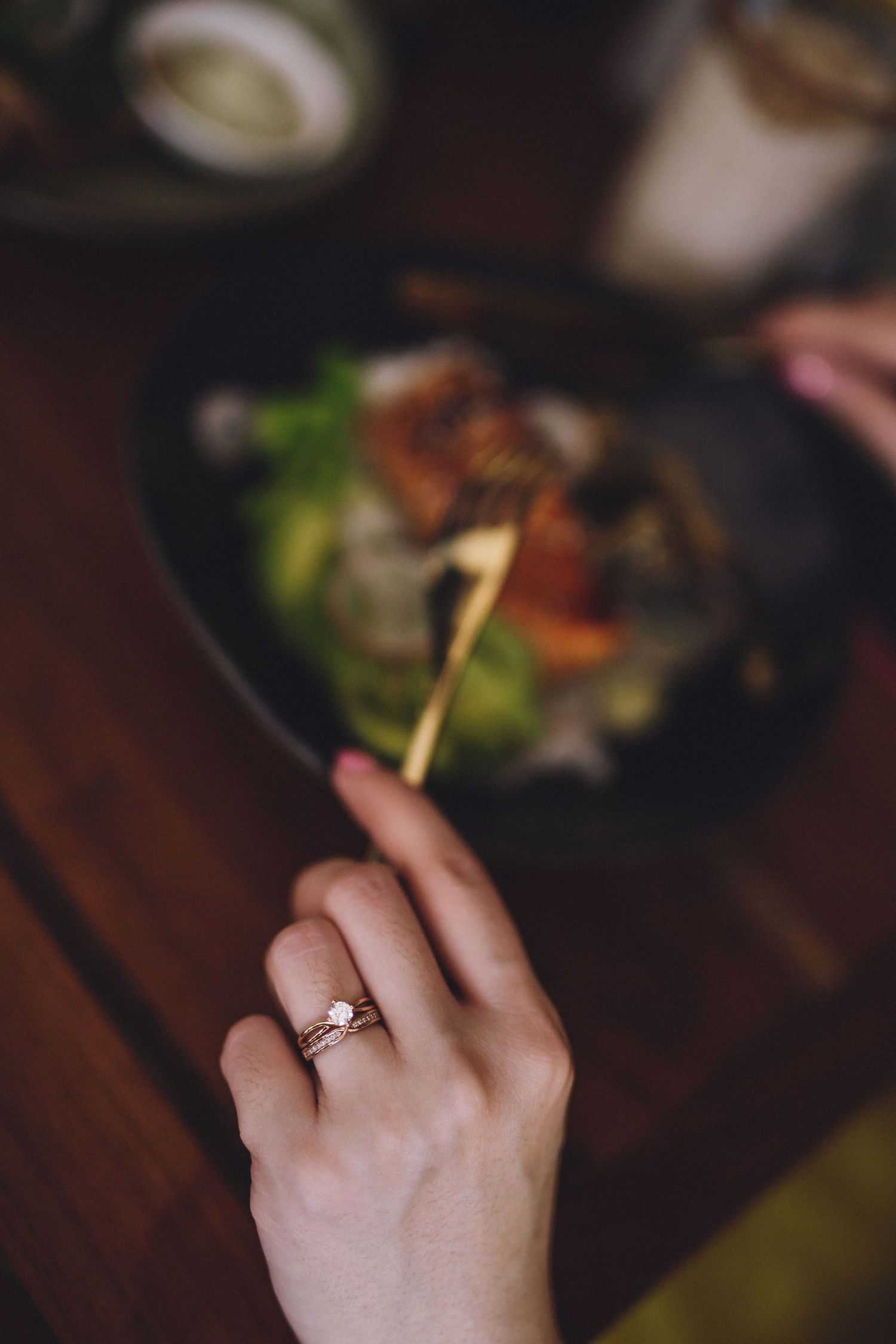 Close-up of a hand wearing an engagement ring with food plates in the background.