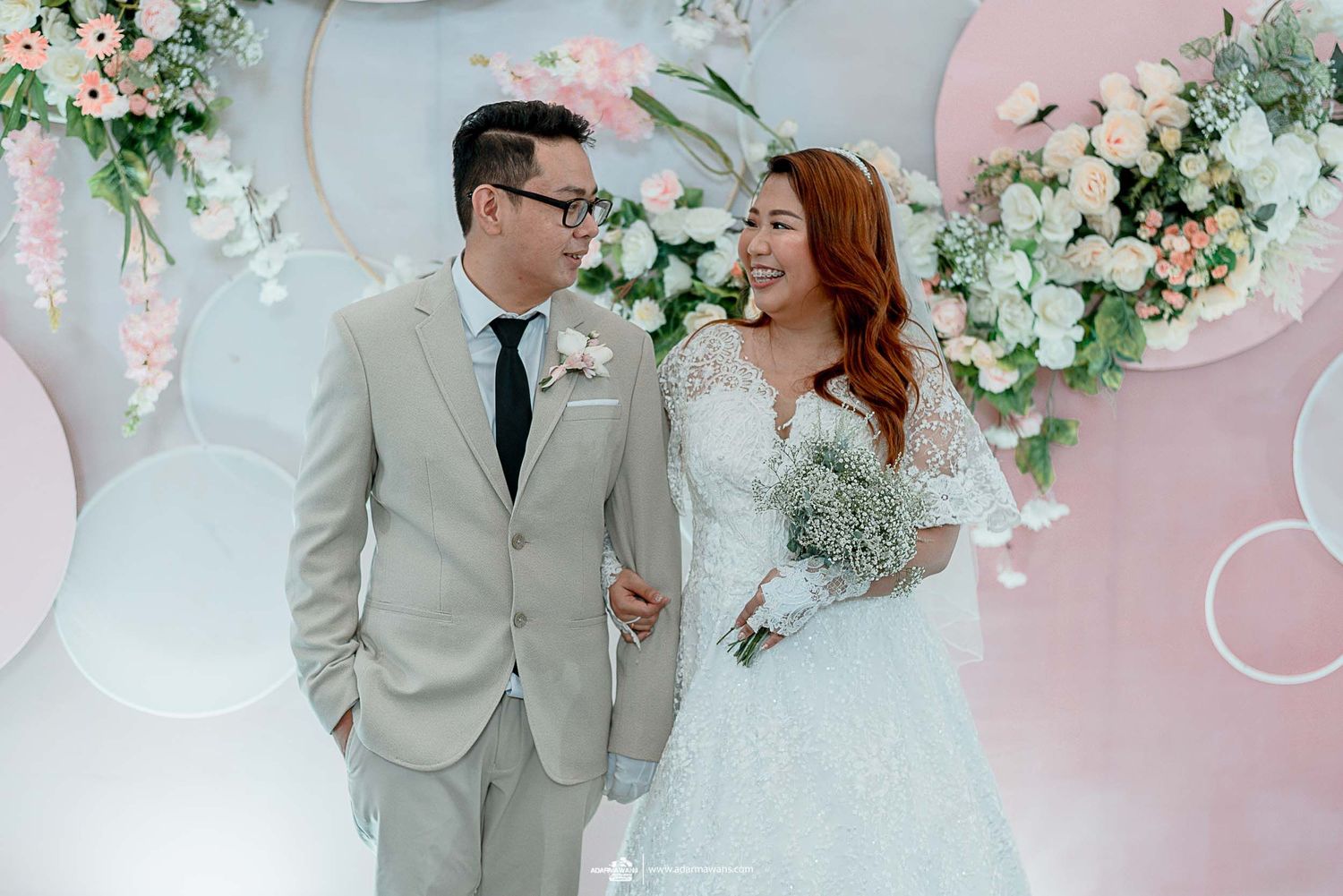 Wedding couple shares joyful moment in front of pink and white floral wall backdrop.