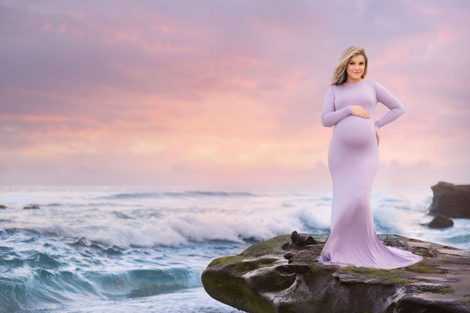 A maternity photo of a woman in a lavender dress standing on rocks by crashing waves at sunset.