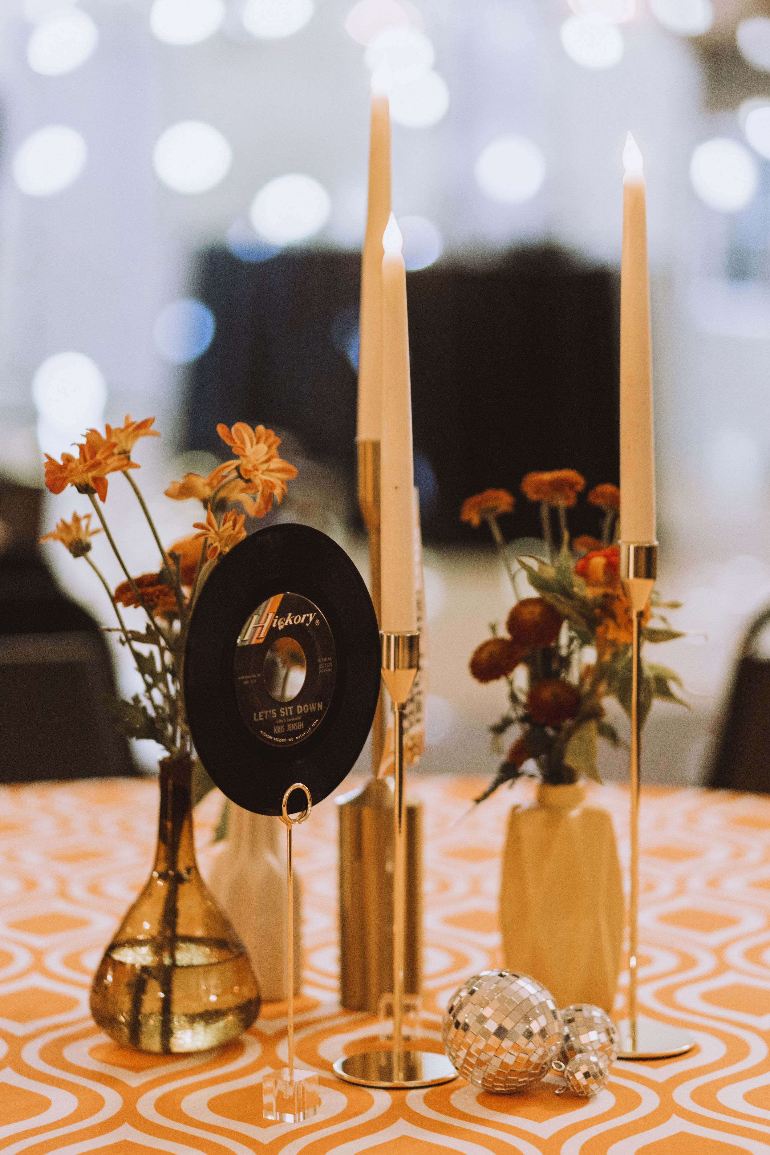 Table centerpiece with vinyl record, orange flowers, and tall cream candles on patterned tablecloth.