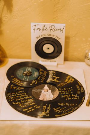 Vinyl records signed with personal messages serve as a unique wedding guest book on a cream-colored surface.
