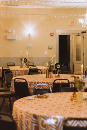 Reception hall setup with round tables covered in orange geometric tablecloths and string lights overhead.