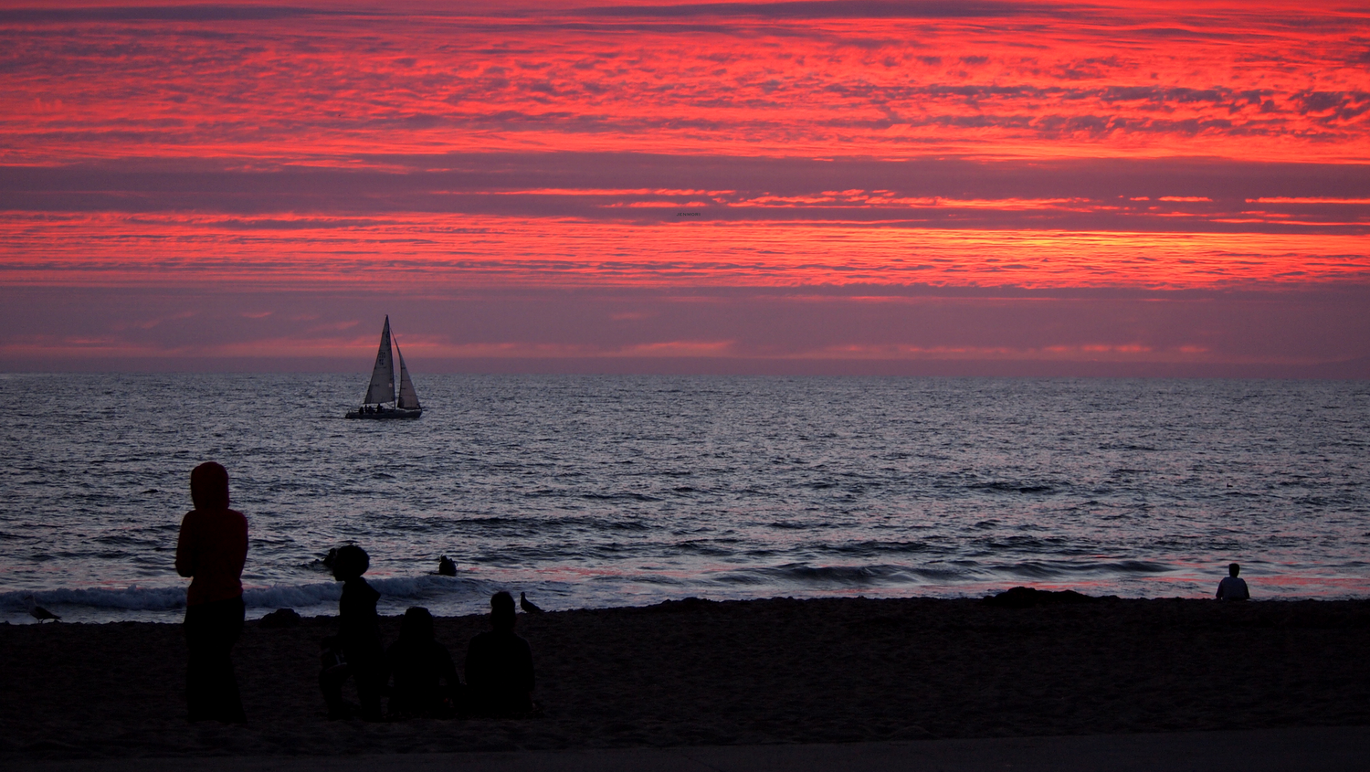 Silhouettes of people on a beach at sunset with a sailboat on the horizon under a vivid red and pink sky.