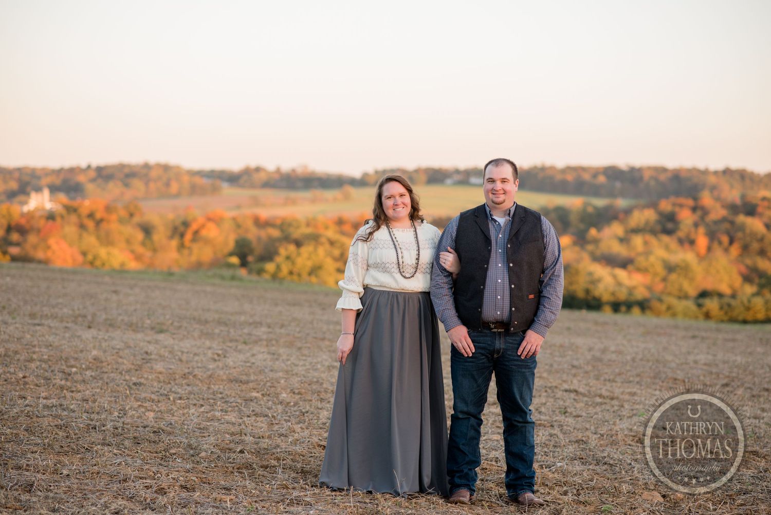 A couple stands together in casual attire against a scenic autumn hillside backdrop at sunset.