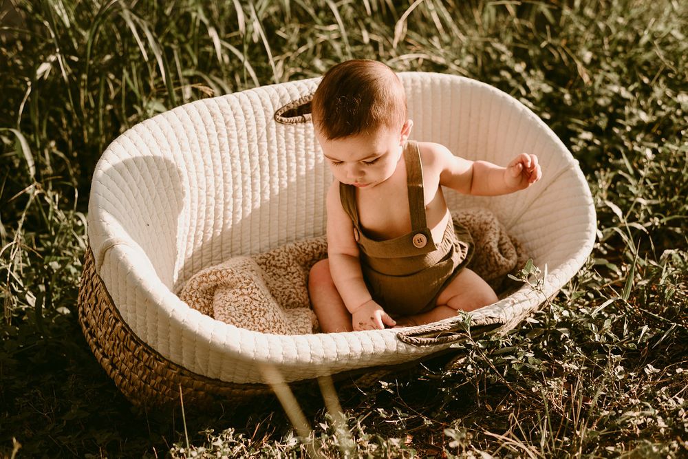 Baby sits in round white woven bassinet placed in tall grass during sunset.