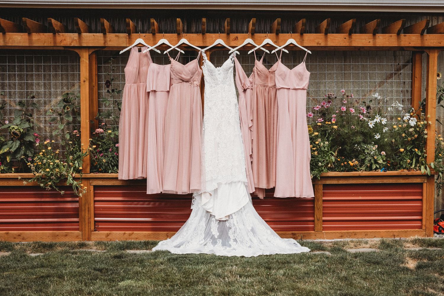 White wedding dress and pink bridesmaid dresses hanging on wooden rack against garden backdrop.