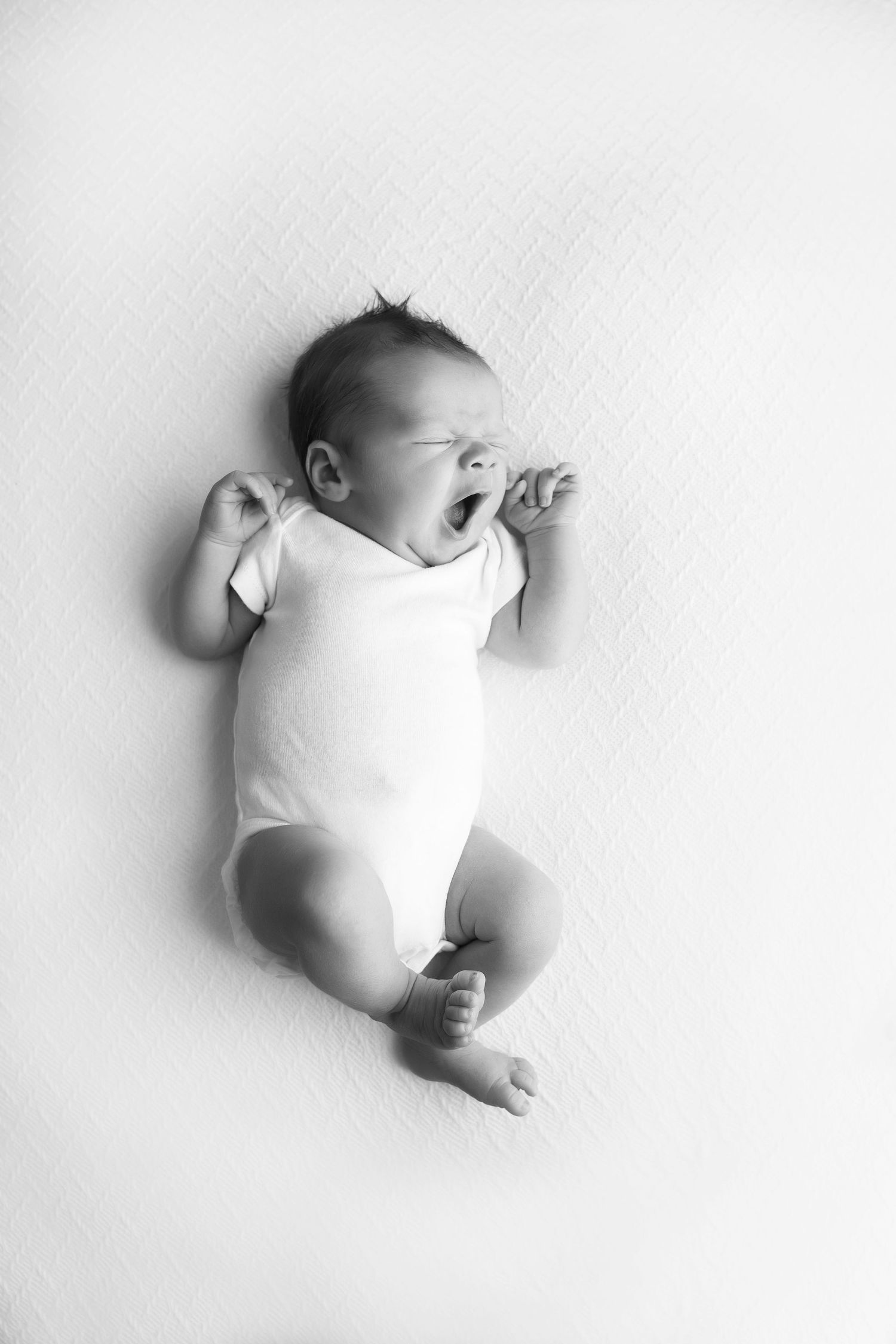 Newborn baby boy laying on white blanket with white onesie.  He is yawning.