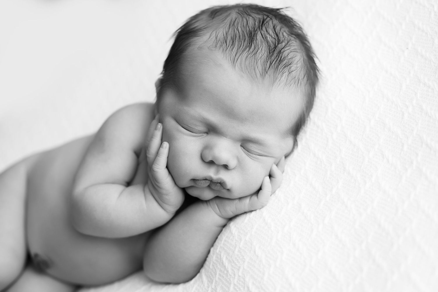 Newborn baby boy on knitted white blanket.  This is a close up of baby's hands hold his cheeks.