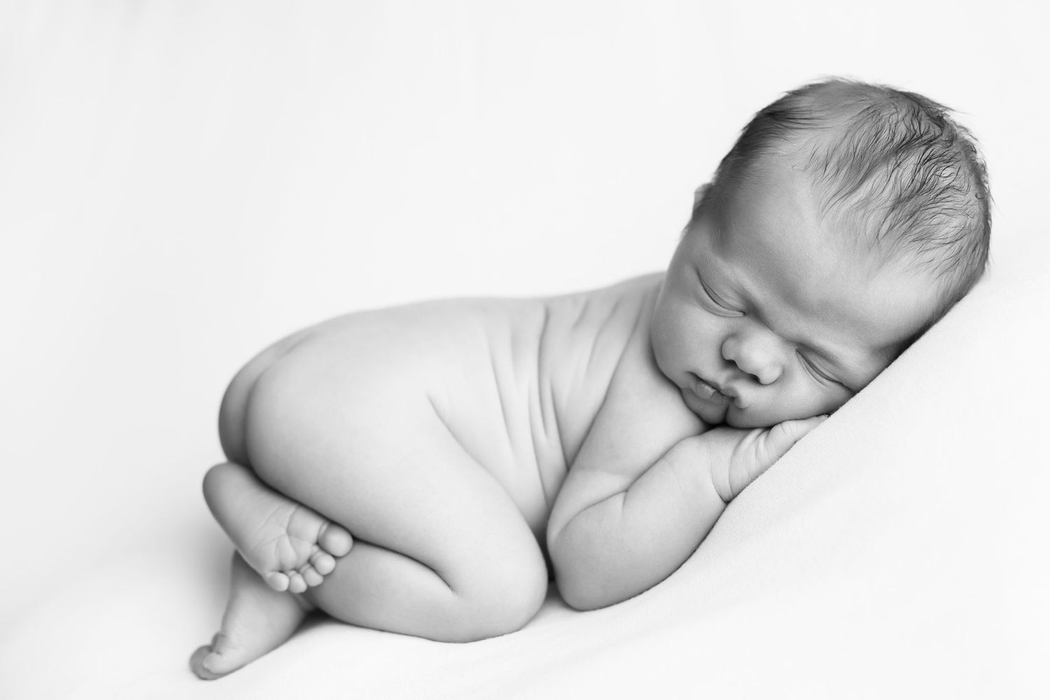 Newborn baby boy on a white blanket.  Baby in on his knees with his hand under his cheek facing towards the camera.