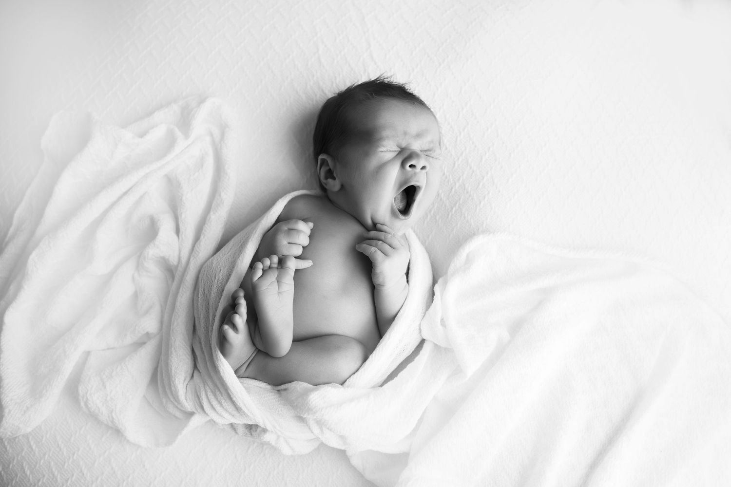 Newborn born baby boy on a white blanket with a white wrap around him.  He is yawning.