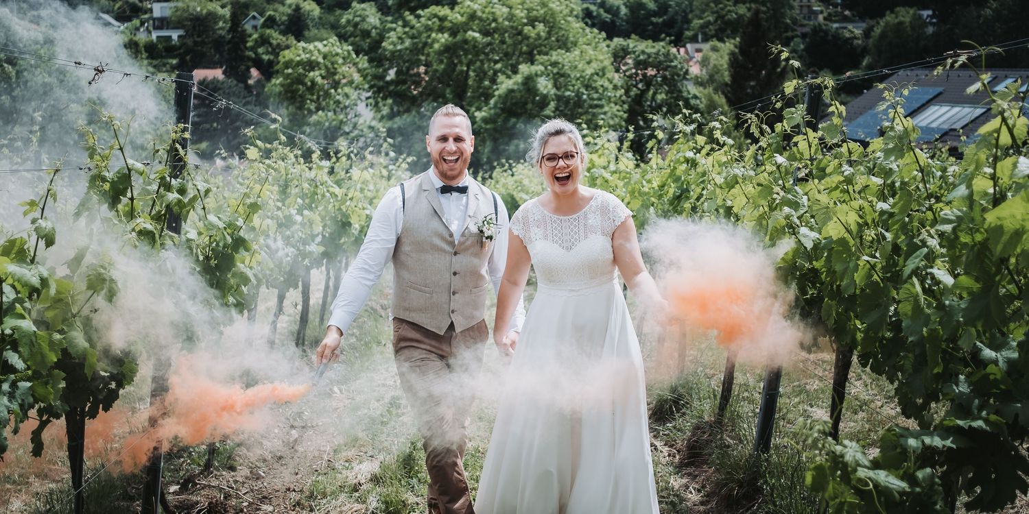 Brautpaar bewegt sich Richtung Kamera des Hochzeitsfotografen mit Rauchfackeln im Weingarten lachend