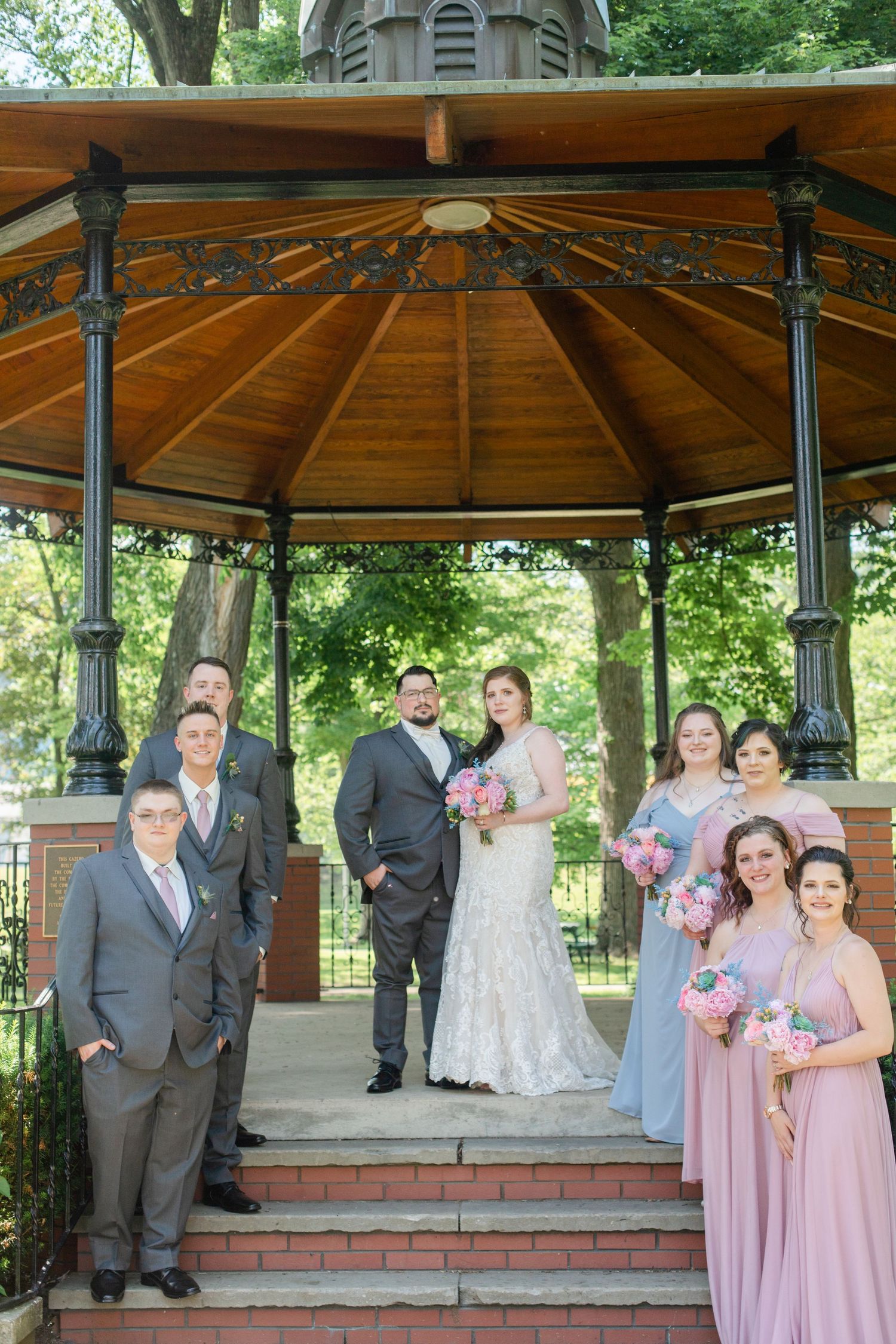 Wedding party poses together in a wooden gazebo with brick steps during outdoor celebration.