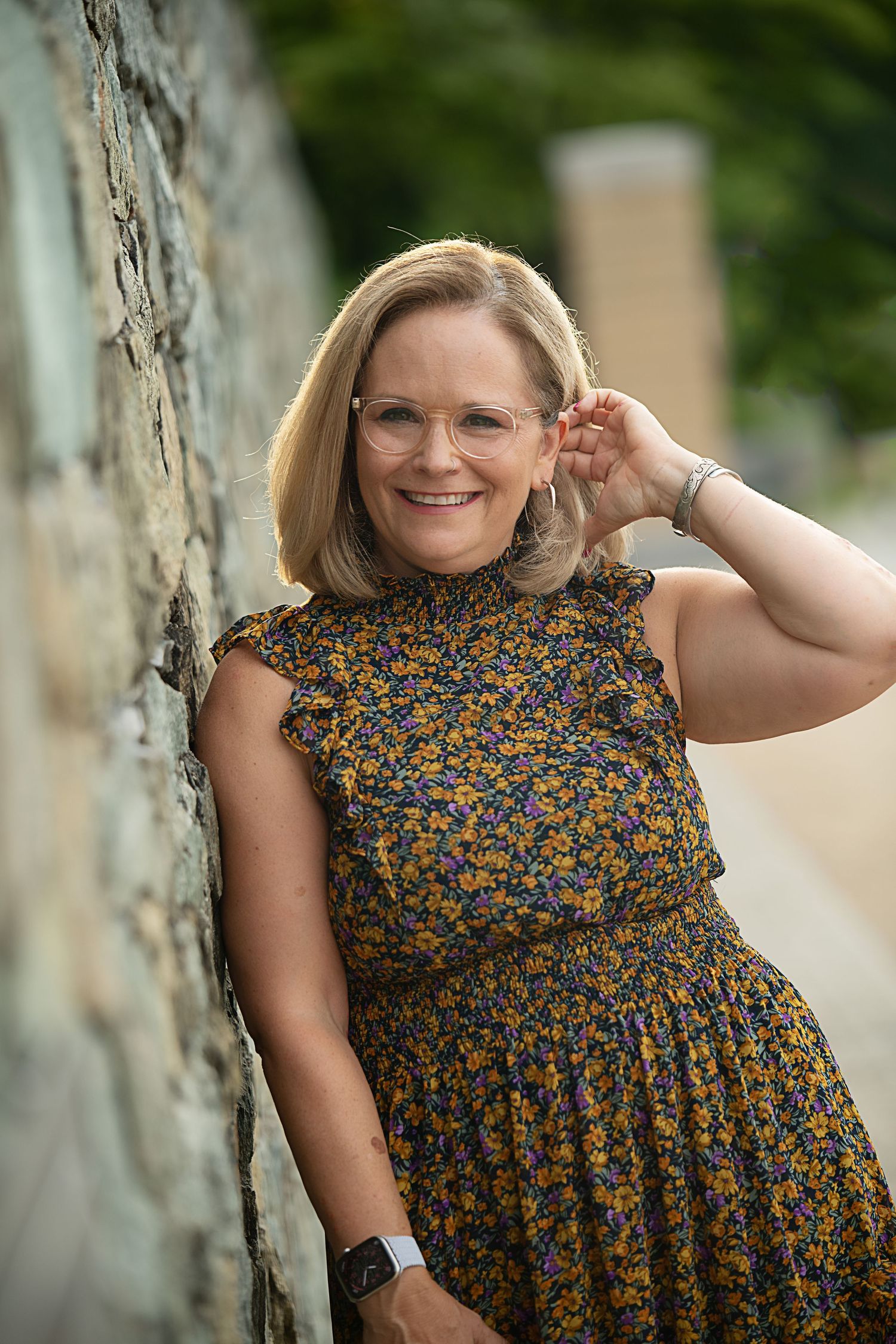A person in a floral dress poses against a stone wall wearing glasses and smiling.