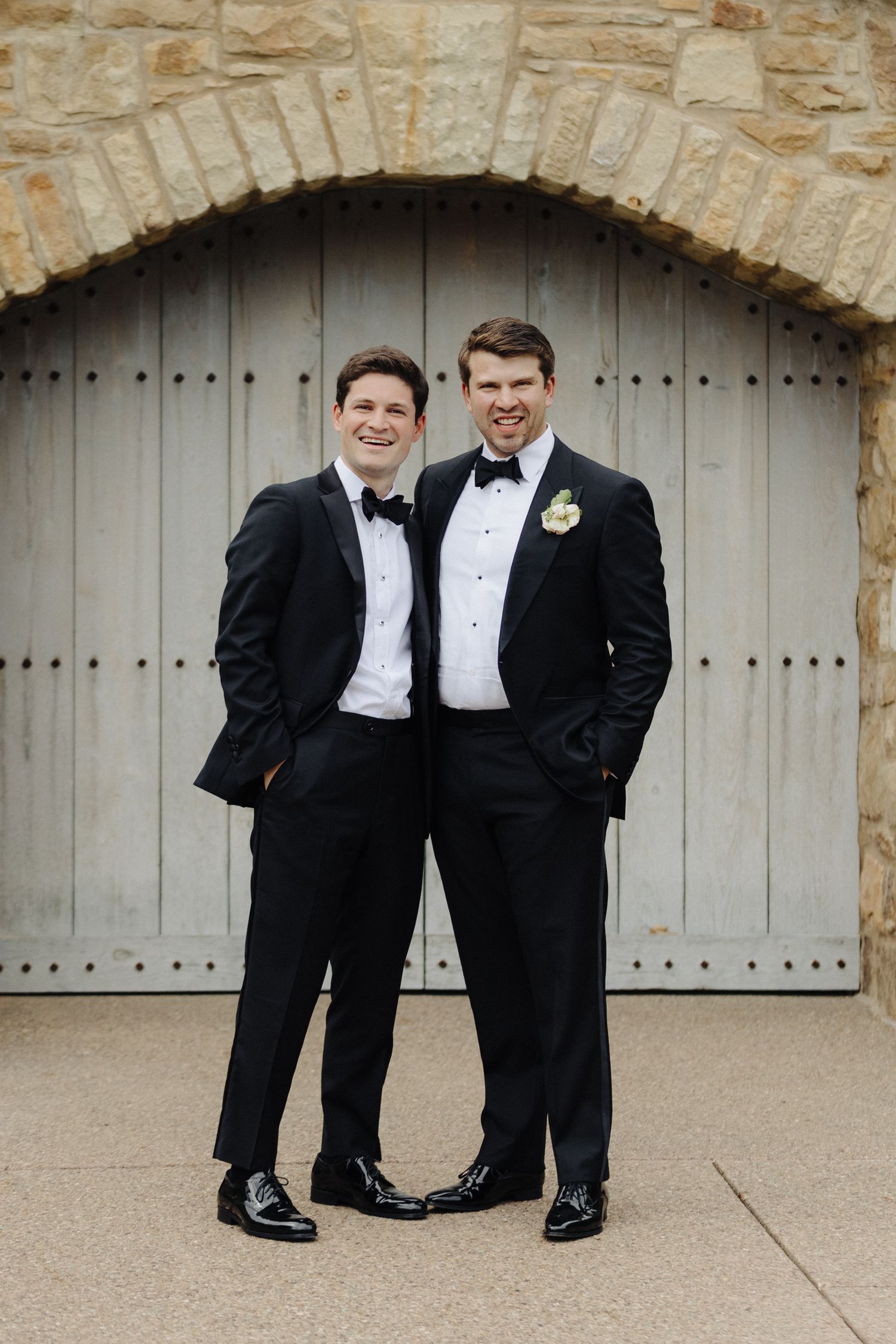 Two men in classic black tuxedos pose in front of rustic stone archway and wooden door.