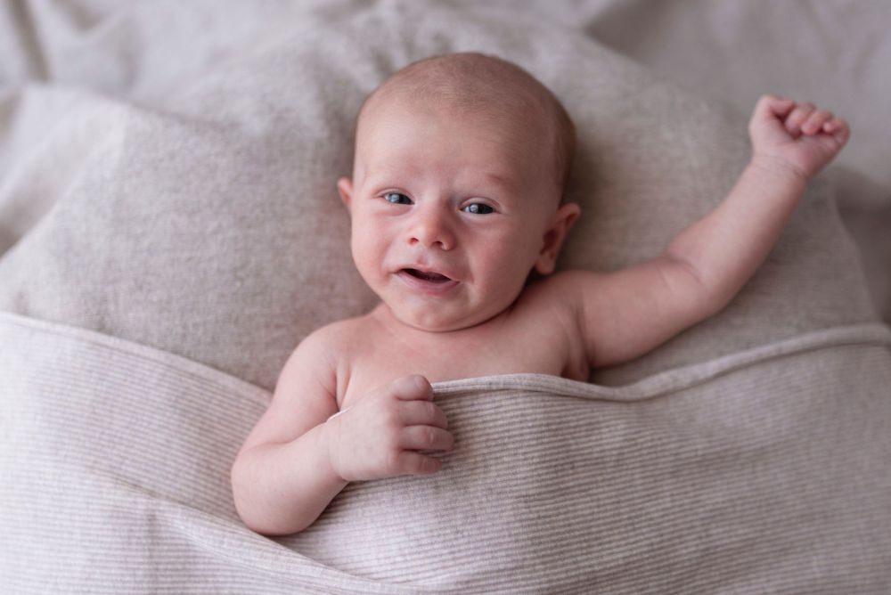 Newborn baby lying on a beige blanket with one arm raised, showing an alert and slightly fussy expression.