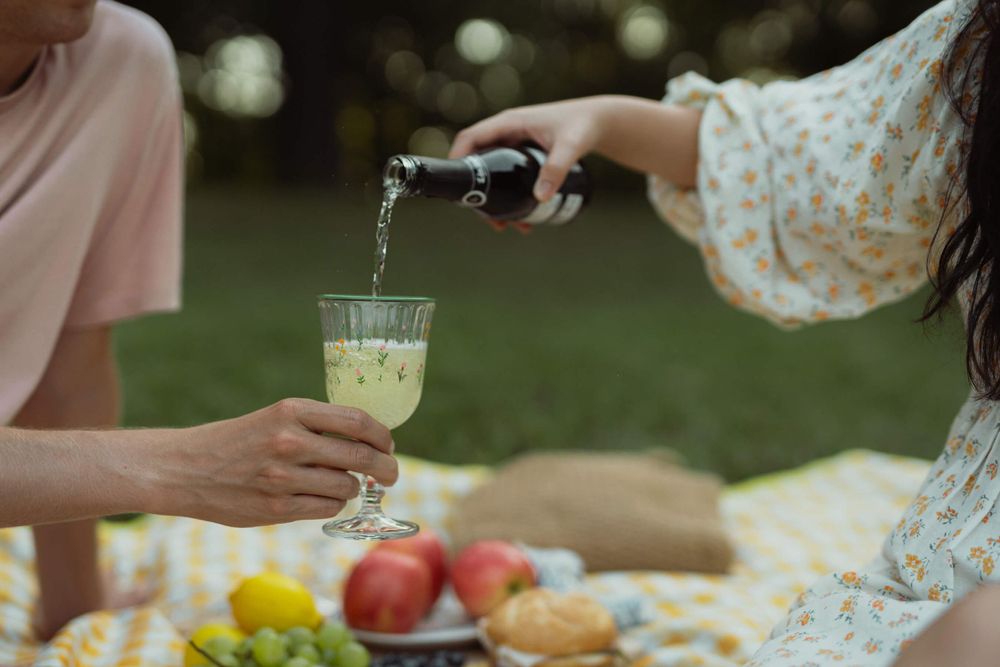 Hands pouring drinks during an outdoor picnic with fresh fruit and snacks.