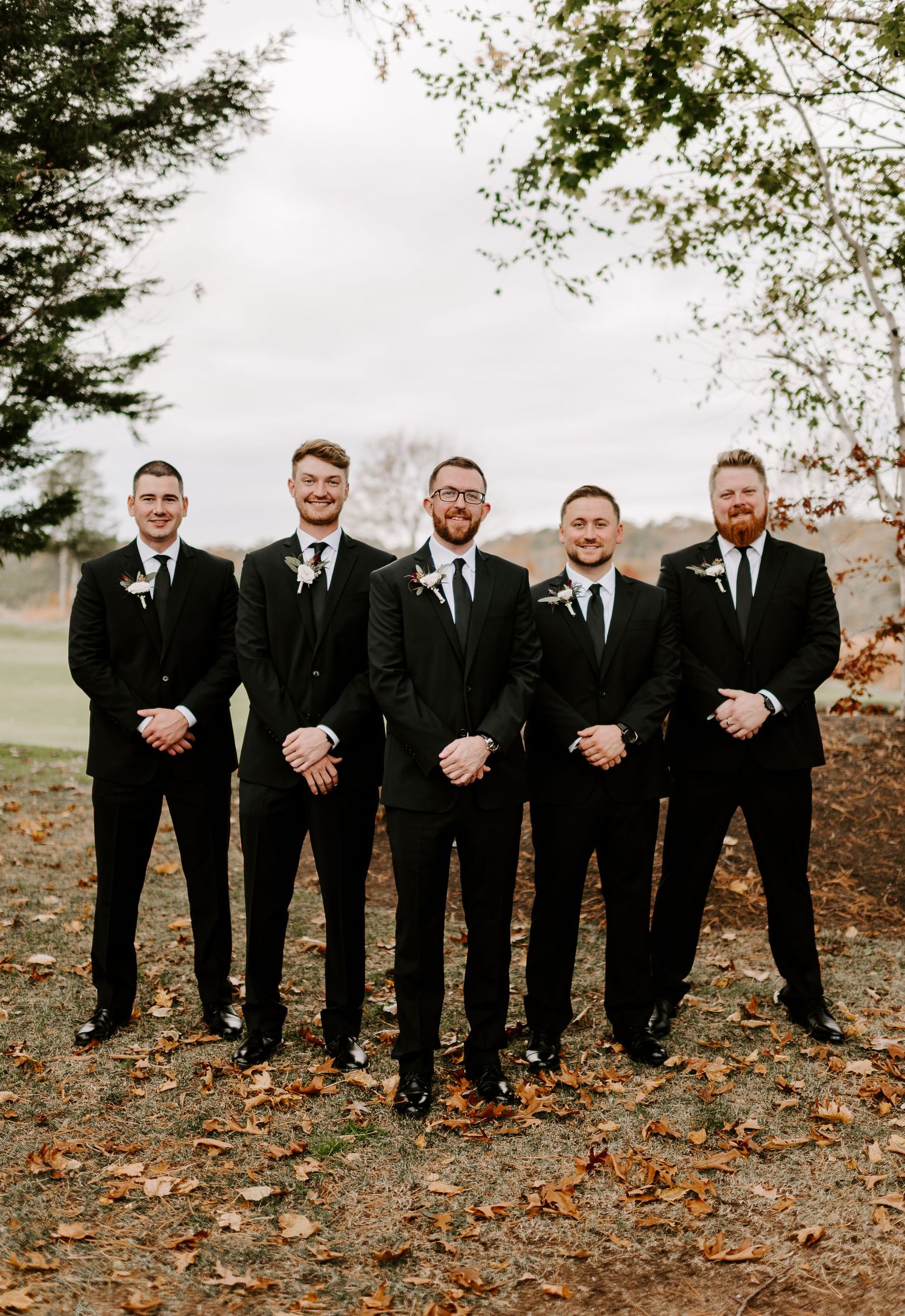 Groomsmen in black suits and ties pose together on autumn day with fallen leaves.