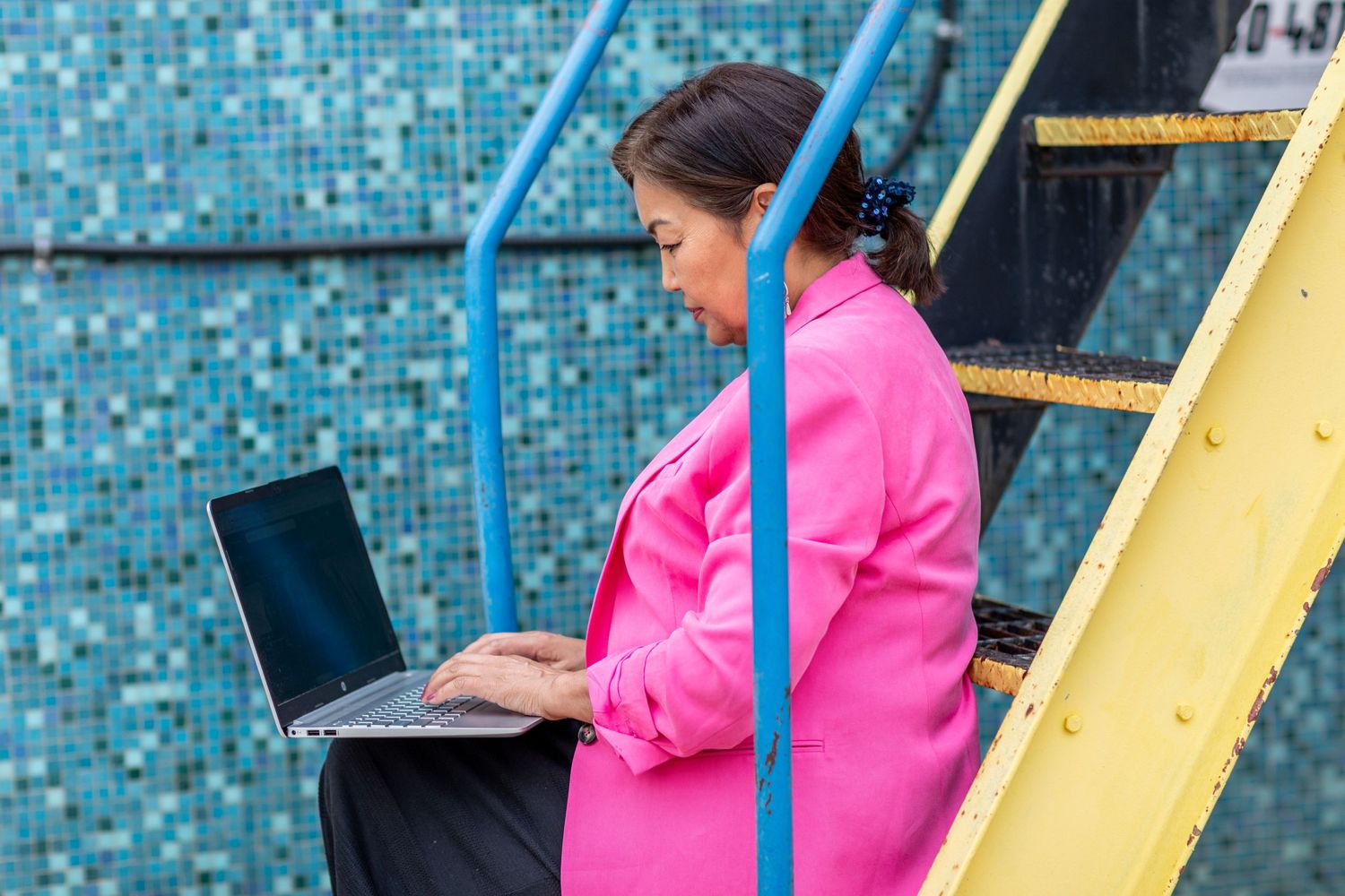 Person in bright pink jacket working on laptop while sitting on stairs with blue railing and mosaic wall background.