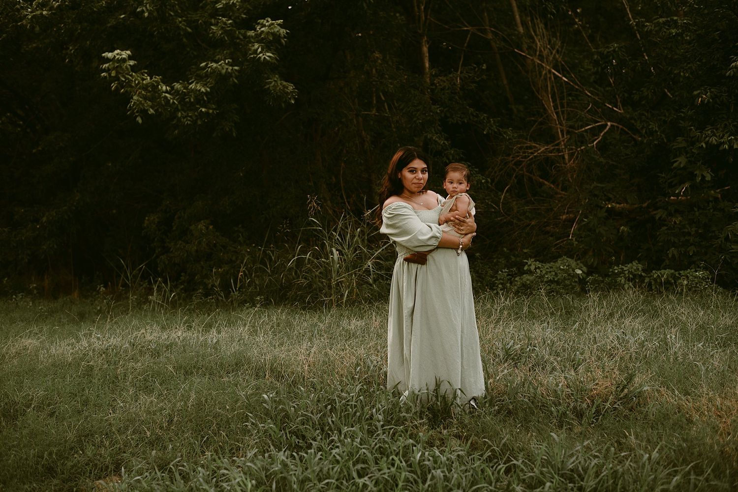A mother wearing a flowing white dress holds a young child in a peaceful outdoor setting at dusk.