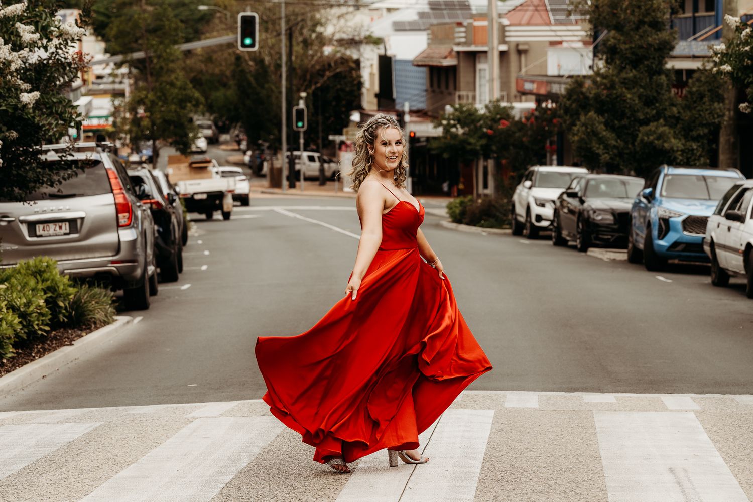 A person in a flowing red dress twirls on a city street with parked cars in the background.