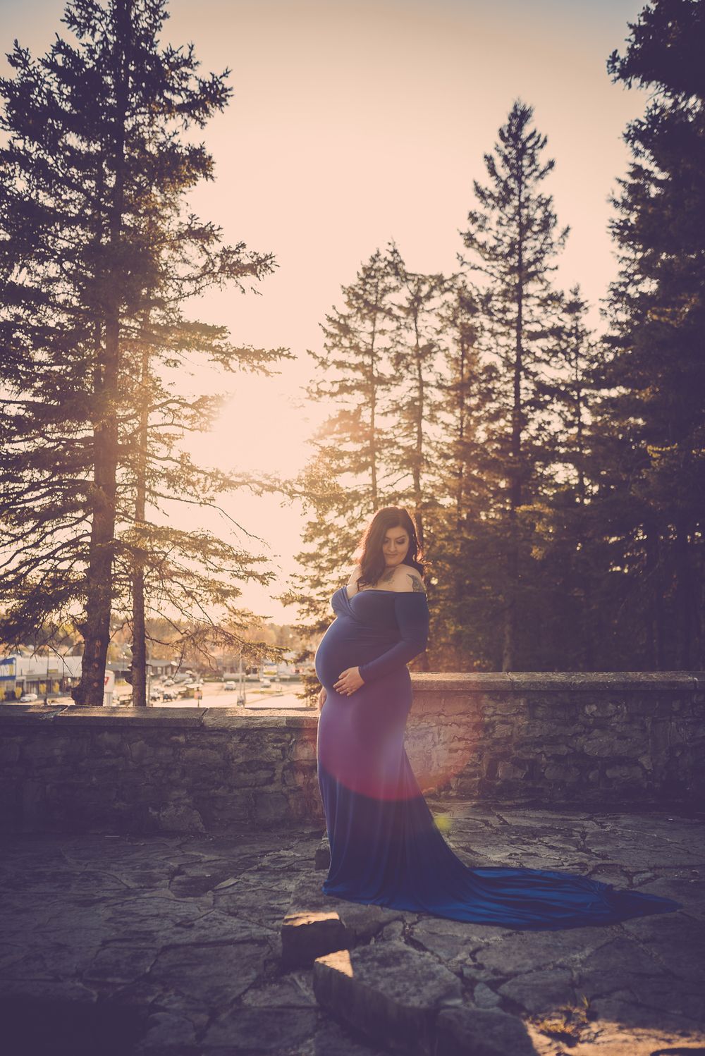 A silhouetted figure in a flowing dress stands against a sunset through pine trees.