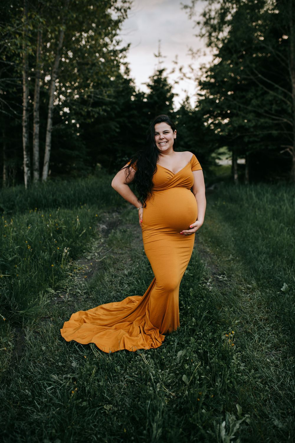 Maternity pose in fitted mustard gown with train on forest path at dusk.