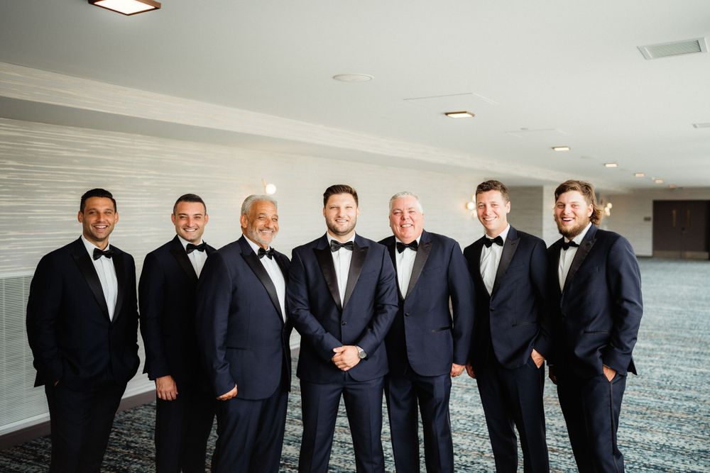 A group of men in black tuxedos and bow ties pose together in a formal wedding party photo.