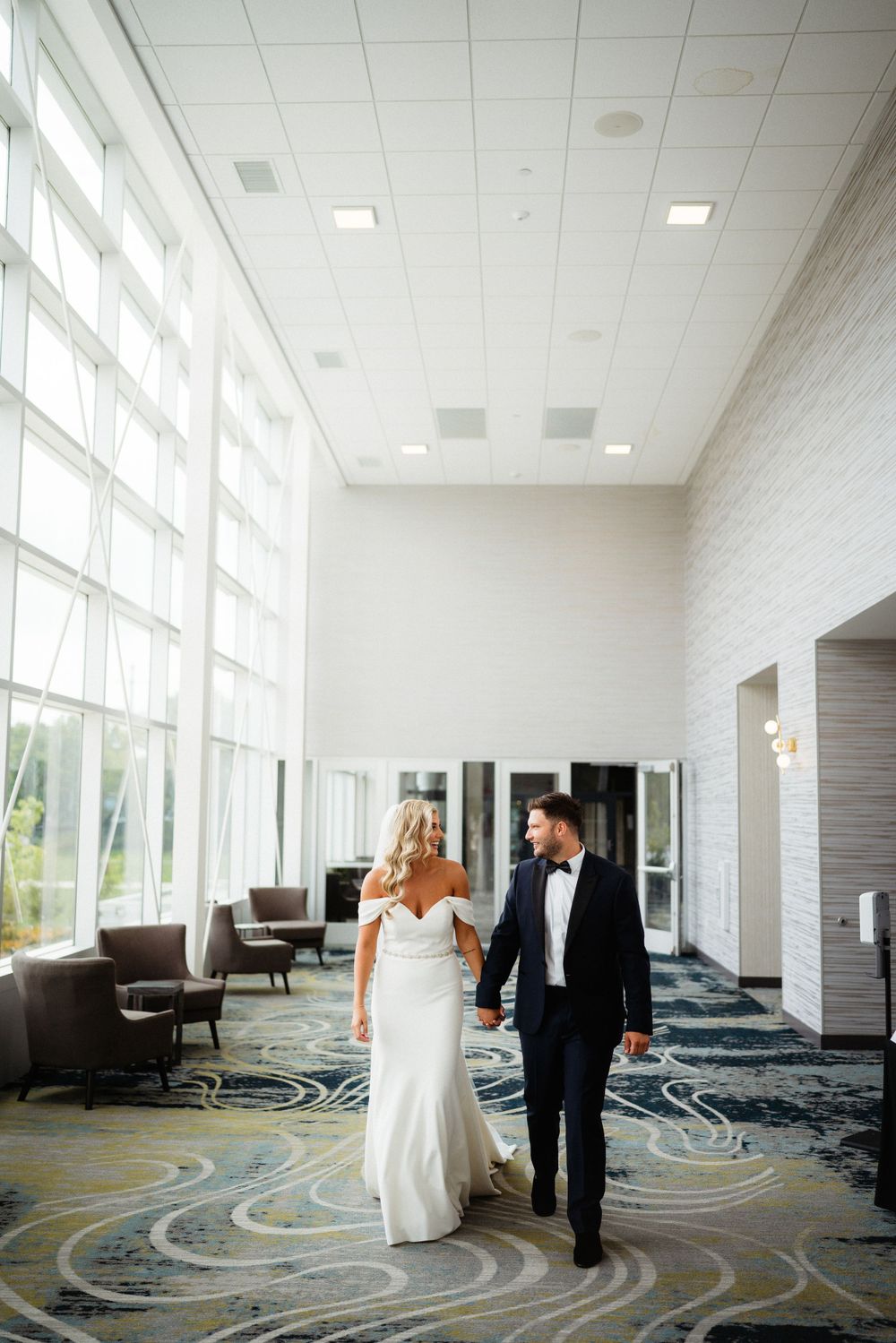 A couple walks together through an elegant hallway with large windows and patterned carpet.
