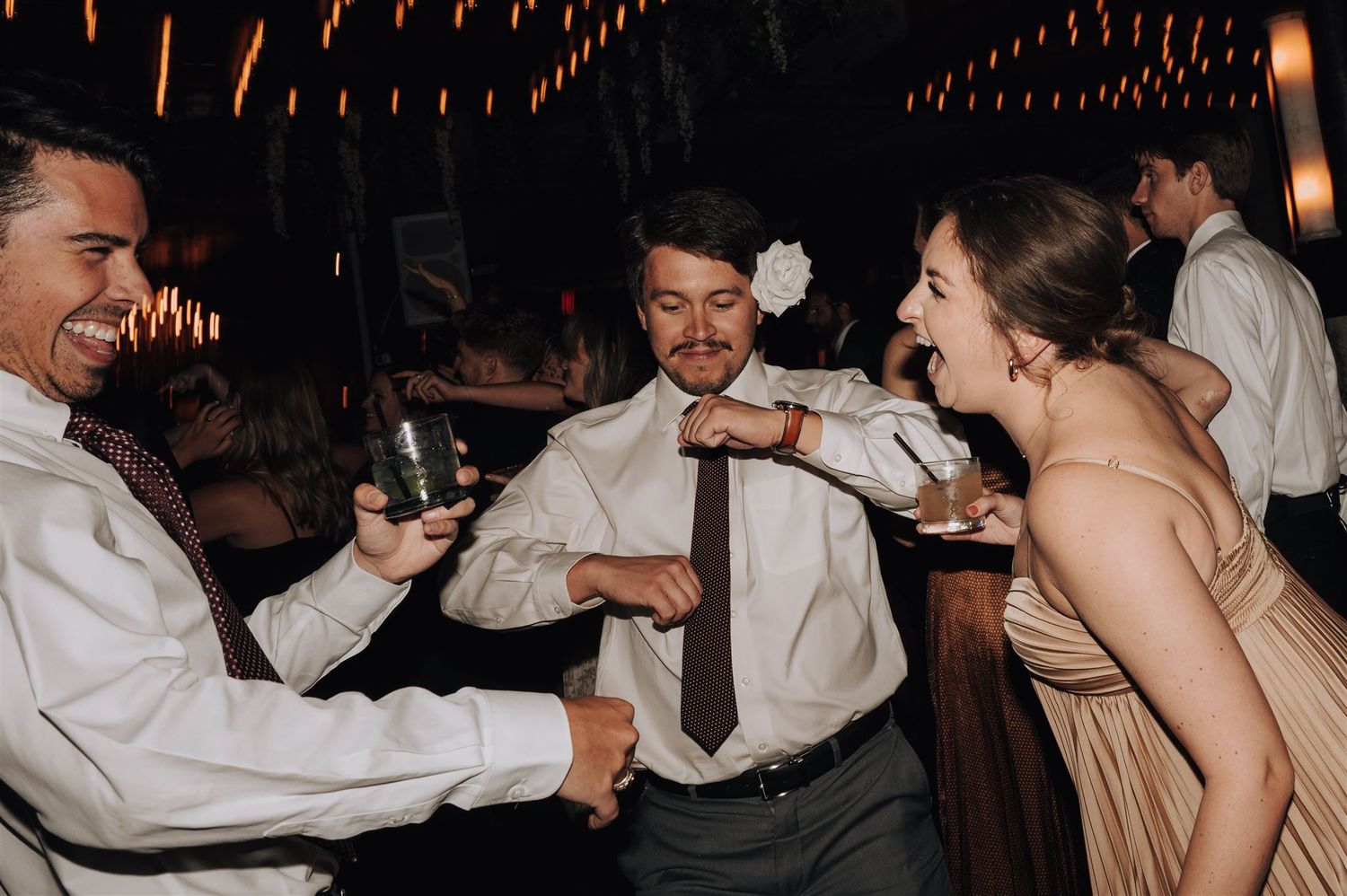 Wedding guests laughing and dancing together under string lights at an evening celebration.
