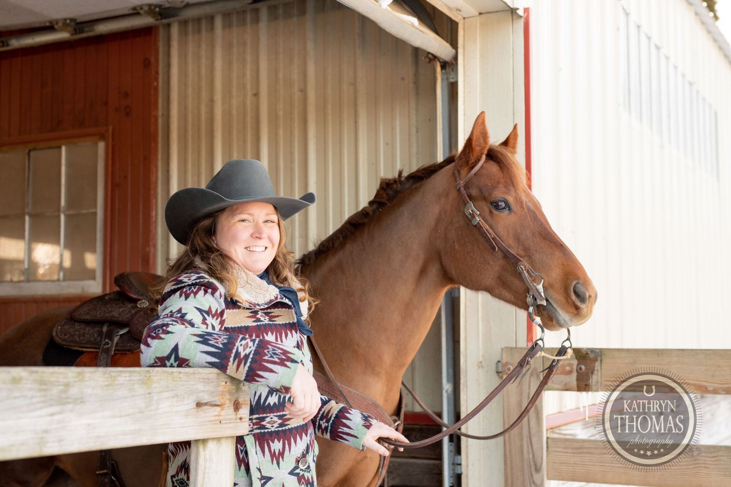 Chestnut horse with bridle stands next to wooden fence at barn entrance.