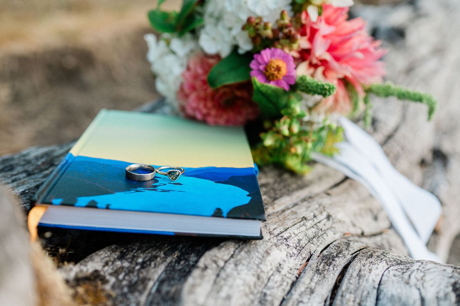 Wedding rings rest on a blue photo album next to a pink and white floral bouquet on a log