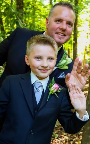 Father and son in matching suits with boutonnieres wave at camera during wedding.