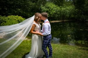Wedding couple shares an intimate moment by a pond with flowing veil in natural setting.