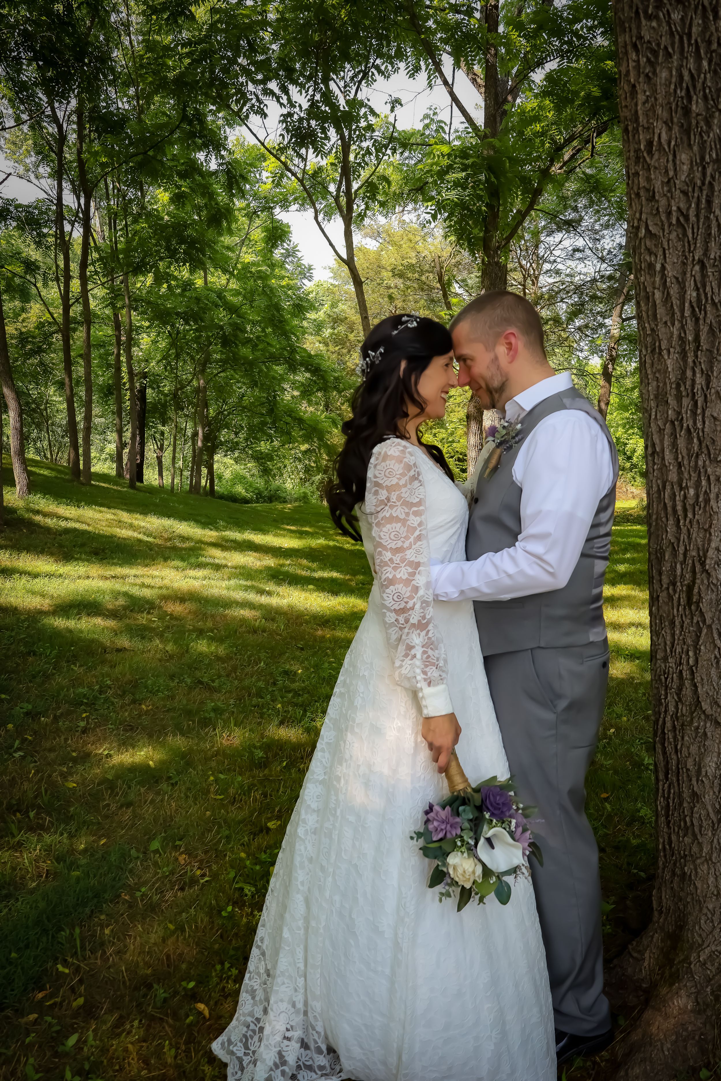 A couple shares an intimate moment among trees during their wedding photoshoot with the bride holding a bouquet.