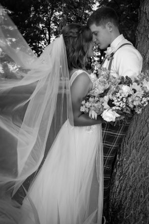 A romantic black and white wedding photo of a couple embracing by a tree with flowing veil and bouquet.
