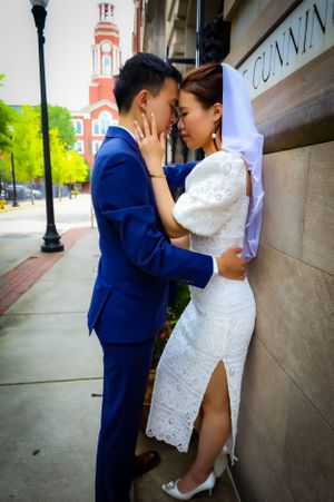 Romantic wedding couple shares intimate moment against brick wall with church spire visible in background.