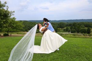 Long wedding veil flows dramatically across green hillside with mountains in background.
