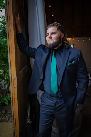 Groomsman in black suit with emerald green tie poses dramatically indoors.