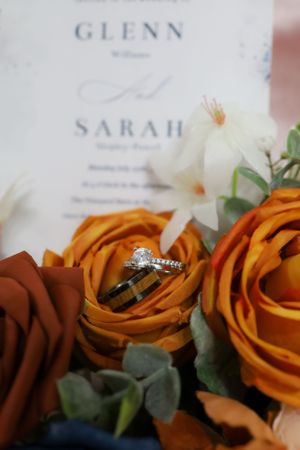 Wedding rings displayed on orange and burgundy roses with invitation cards.