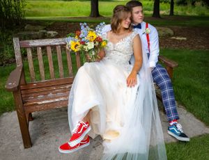 Newlyweds in formal attire and casual sneakers sit on wooden bench with colorful bouquet.