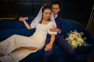 Wedding couple relaxes on blue couch with bridal bouquet in moody lighting.