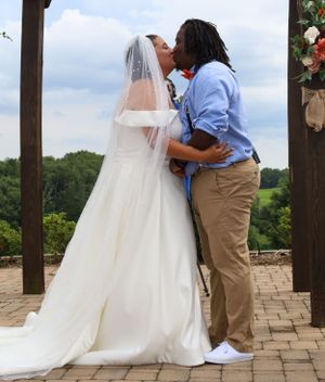 Wedding ceremony takes place under wooden pergola on stone patio with floral decorations.