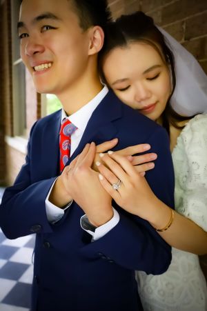 Newlywed couple embraces in romantic moment showing off wedding ring against brick wall backdrop.